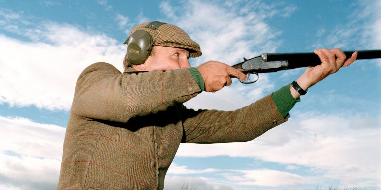 A man in tweed shooting a shotgun against a sky