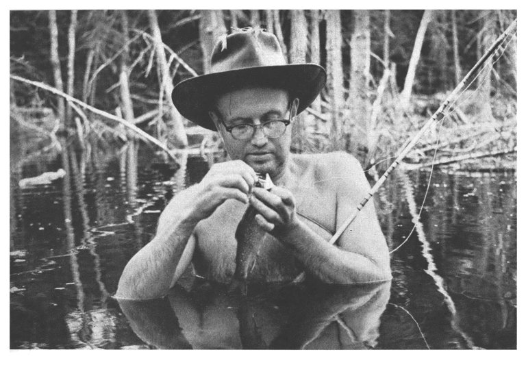 A black and white photo of a man chest deep in a bog looking at a fishing fly.