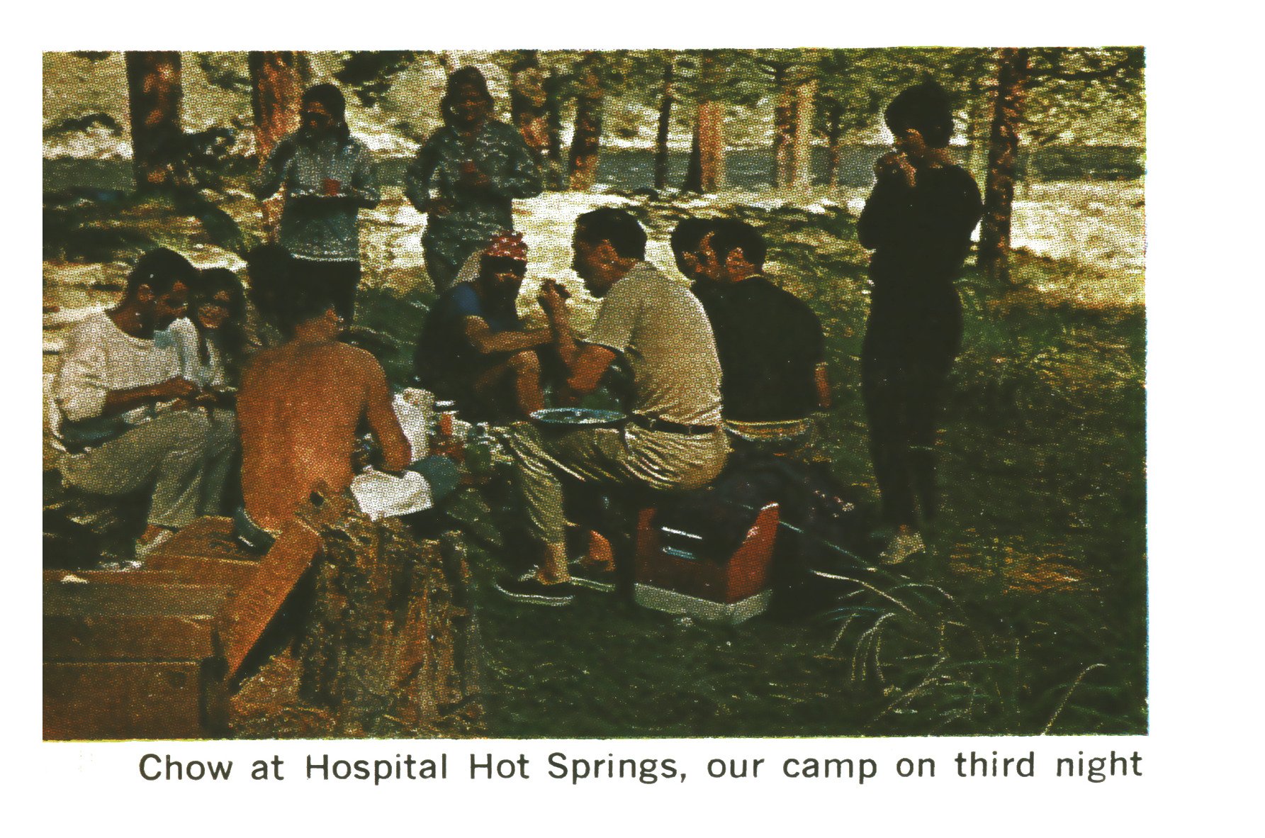 Trip members sit and eat around a riverside camp in a pine forest, with a cooler and gear box in the foreground.