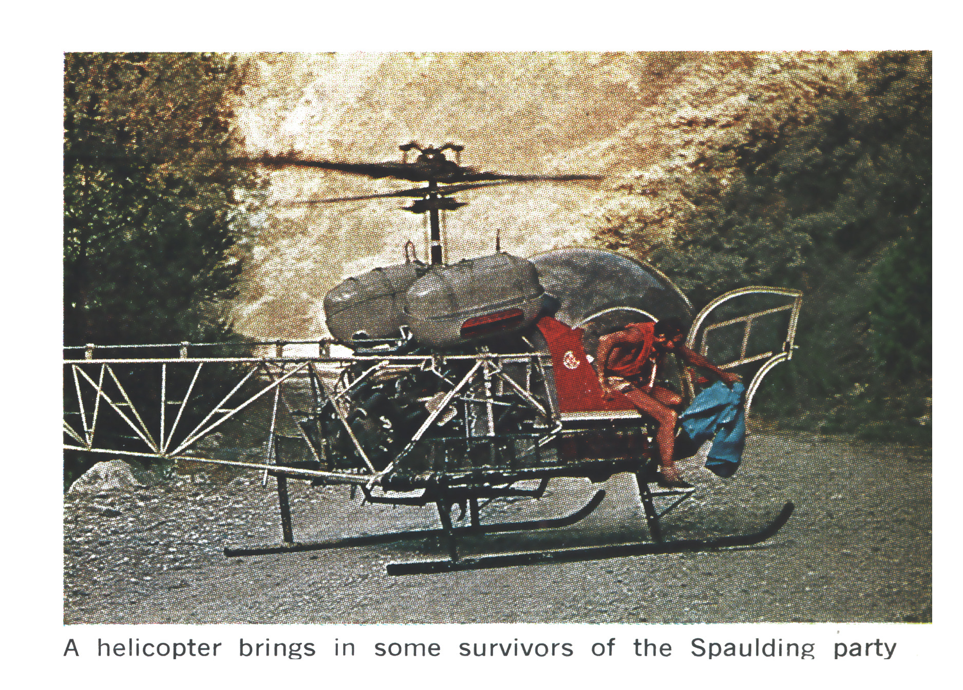 A small bubble-canopy helicopter with a person in red rests on a gravel road beside a wooded riverbank.