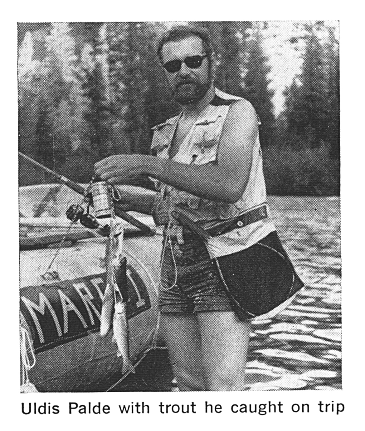 Black-and-white photo of a man in a vest and shorts standing beside a raft marked "MARK I," holding a fishing rod and small trout.