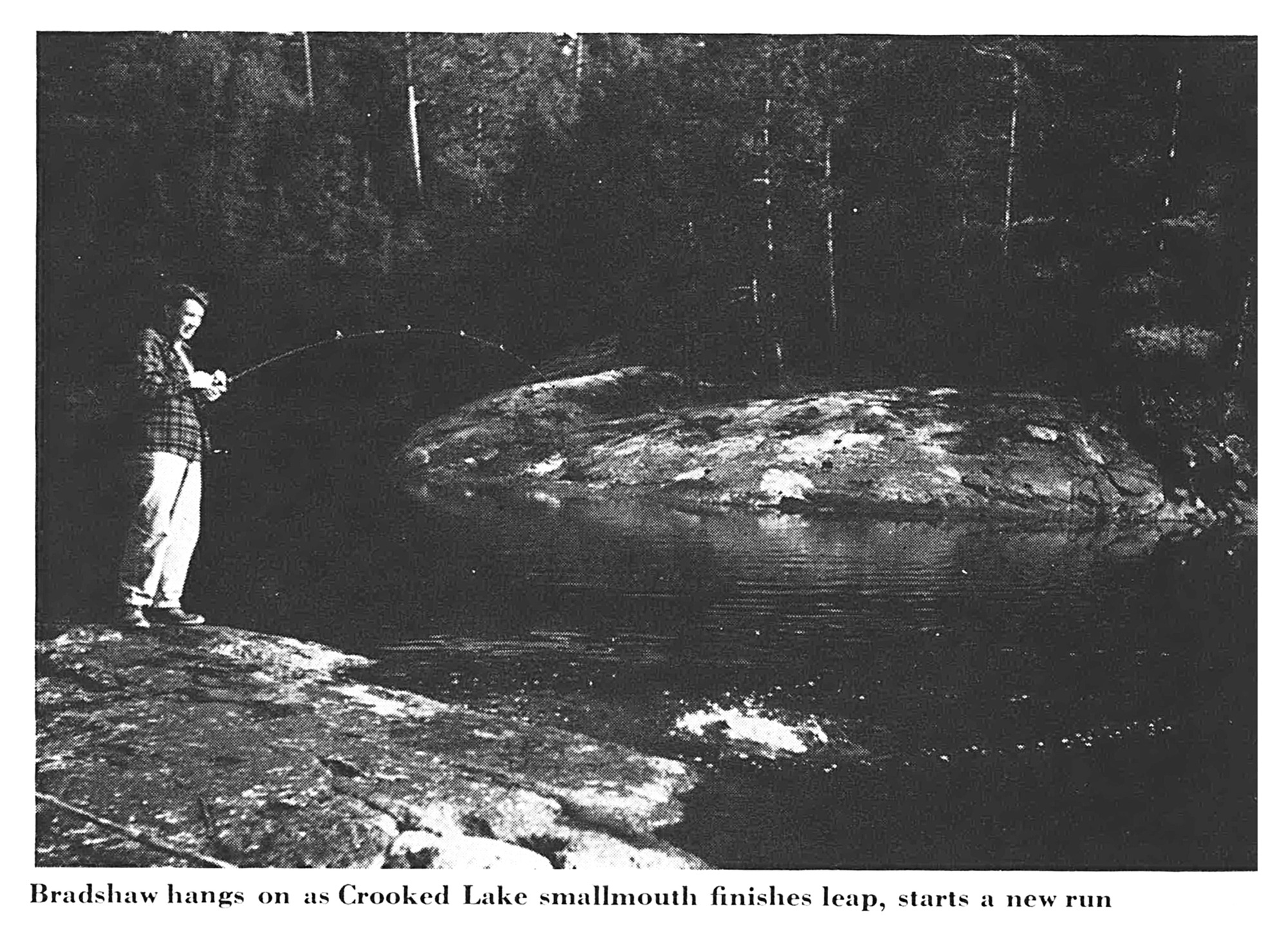 An angler braces on a shoreline rock as a smallmouth finishes a leap and starts a new run (black-and-white photo).