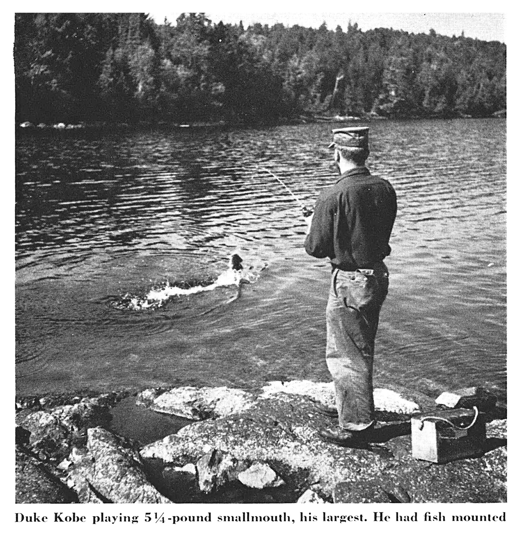 Guide Duke Kobe stands on a rock ledge, rod bent, playing a smallmouth bass in the lake (black-and-white photo).