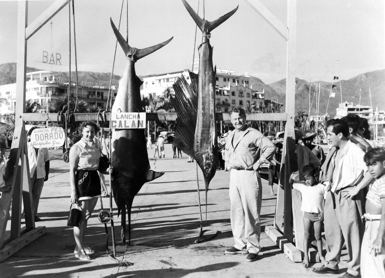 A woman and man pose with their day's catch, a 225 pound black marlin and a sailfish, caught on the fishing boat Calan in waters near Acapulco, Mexico in 1954 as onlookers stand by.