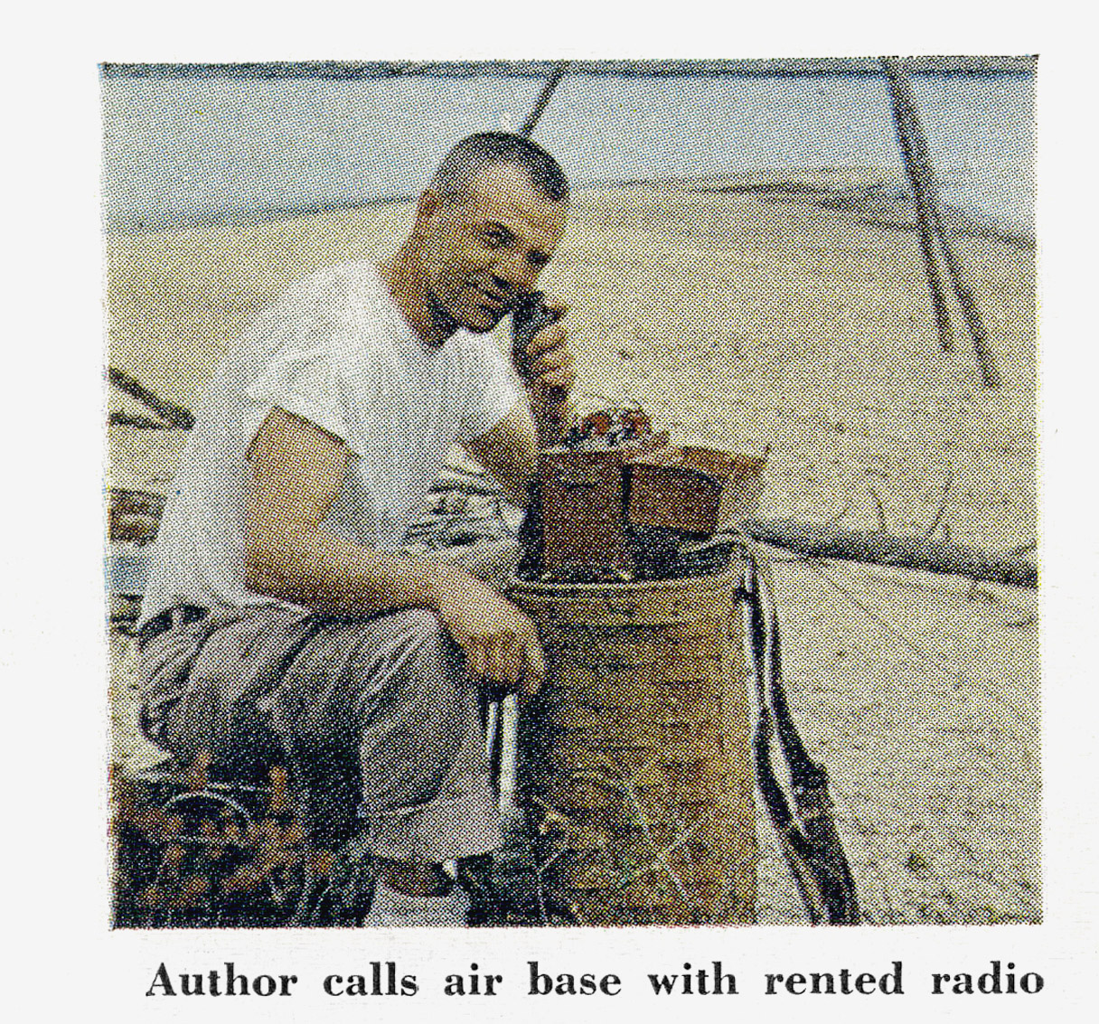 A man kneeling on a beach speaks into a small rented two-way radio set on a packbasket, calling the air base.
