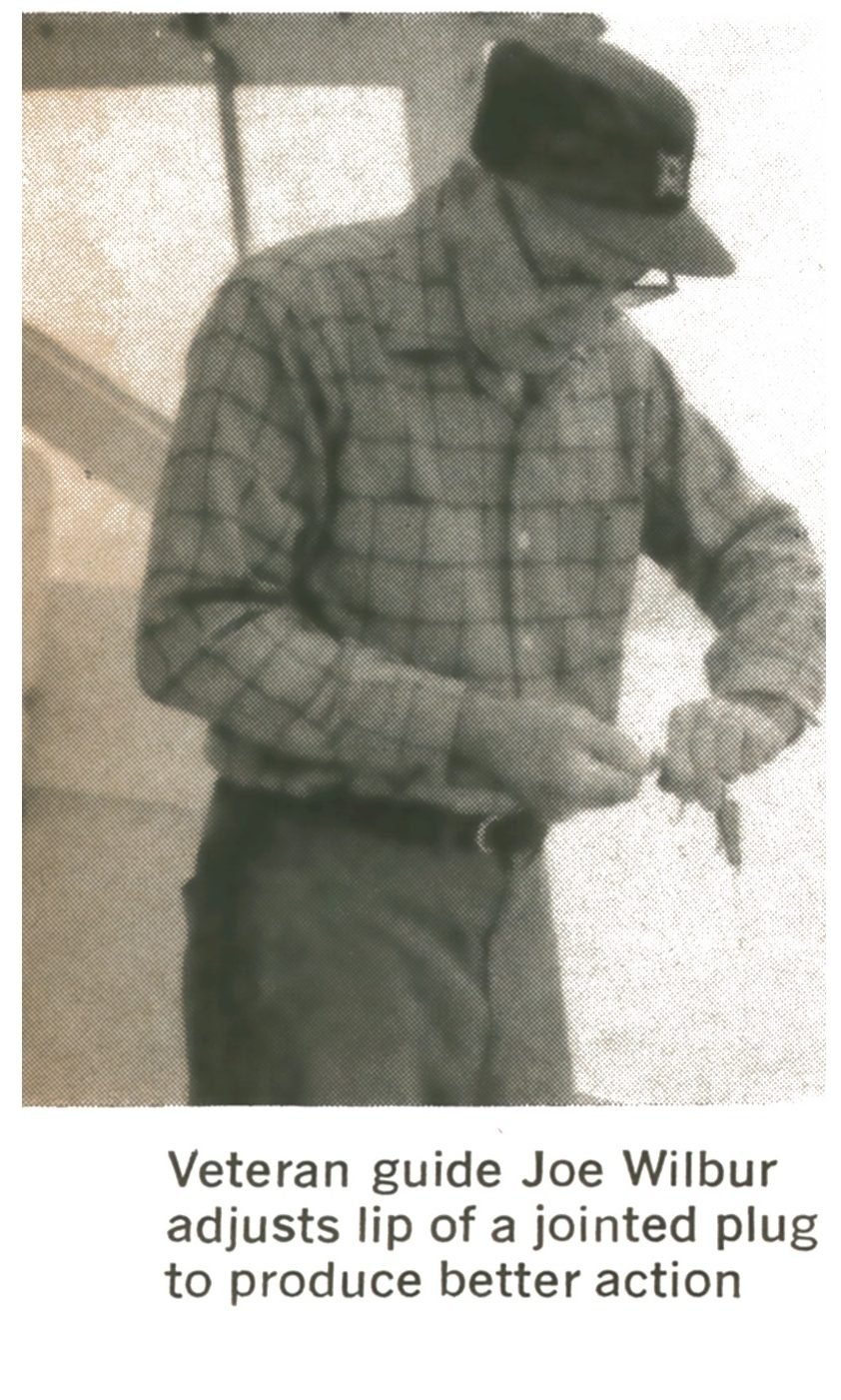 Black-and-white photo of veteran guide Joe Wilbur in a plaid shirt and cap adjusting the lip of a jointed fishing plug.