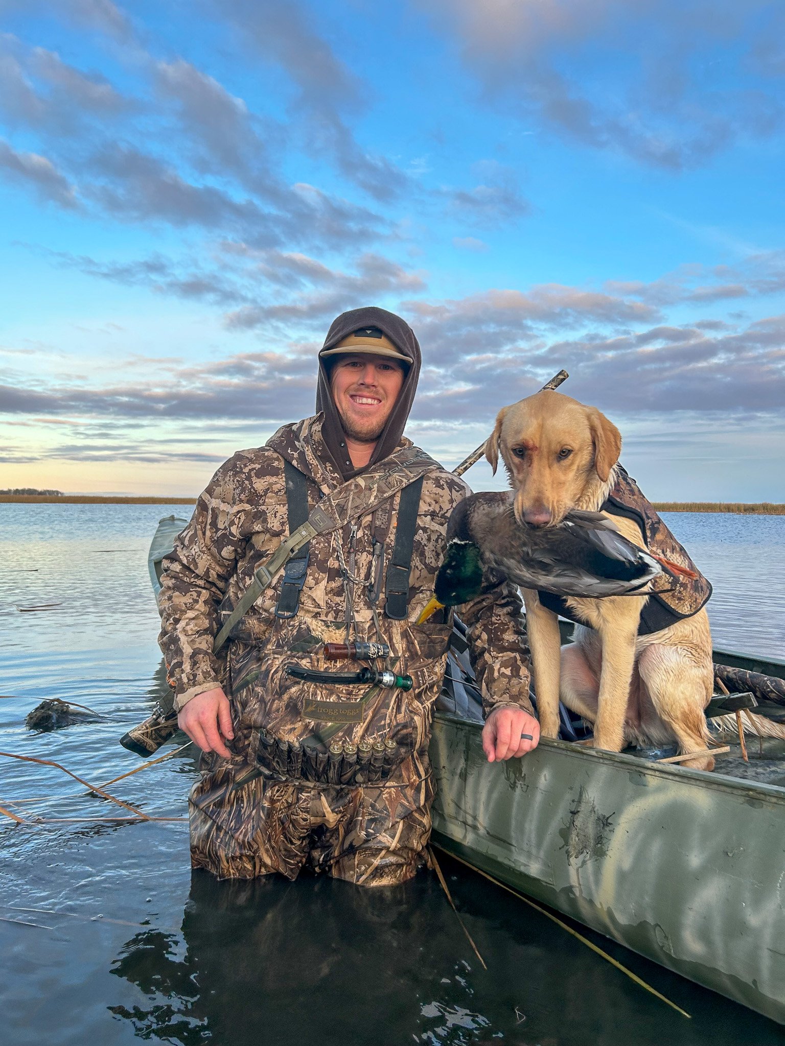 A duck hunter and his dog on a boat, holding a mallard.