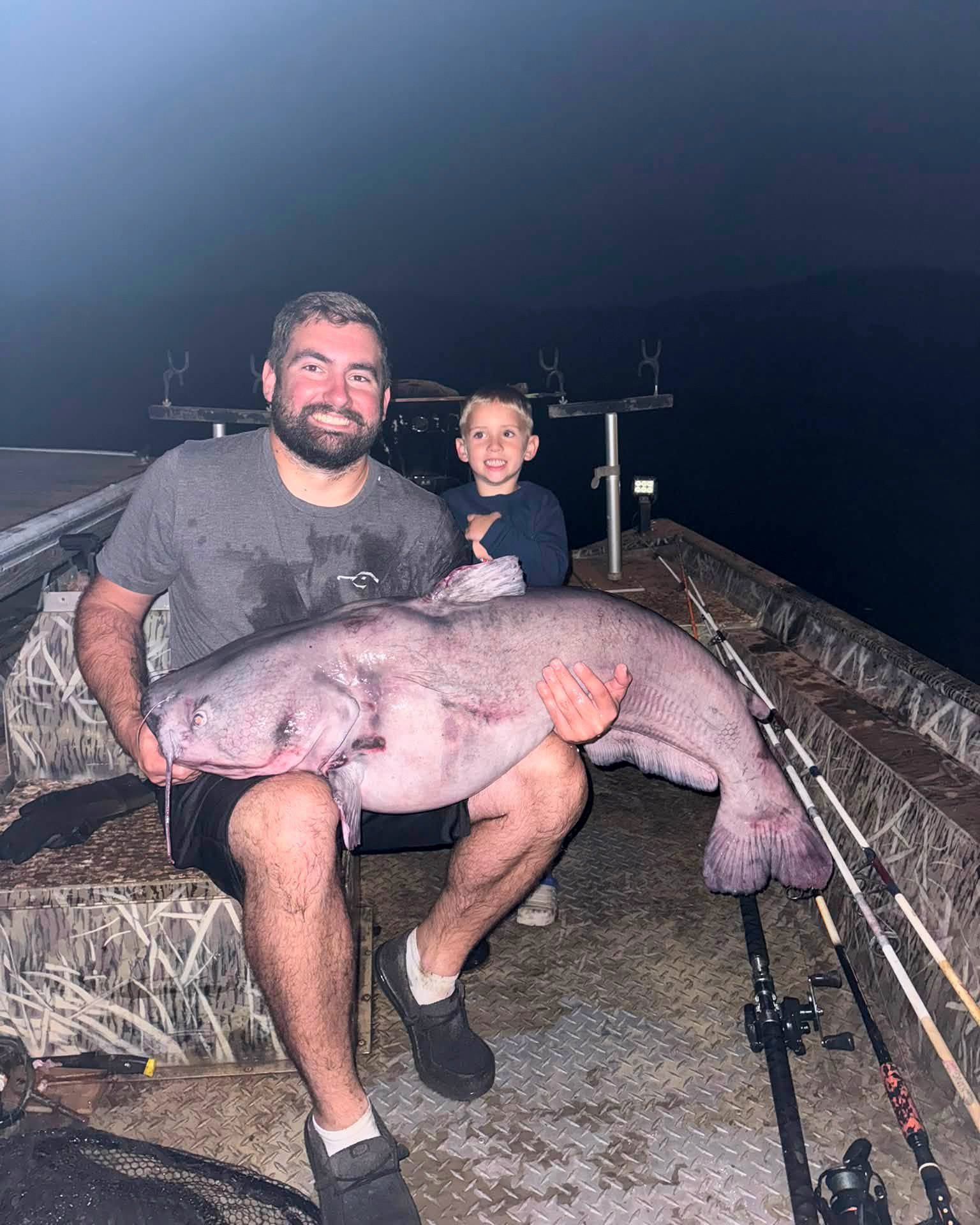 A man holds a big blue catfish while his son sits behind him.
