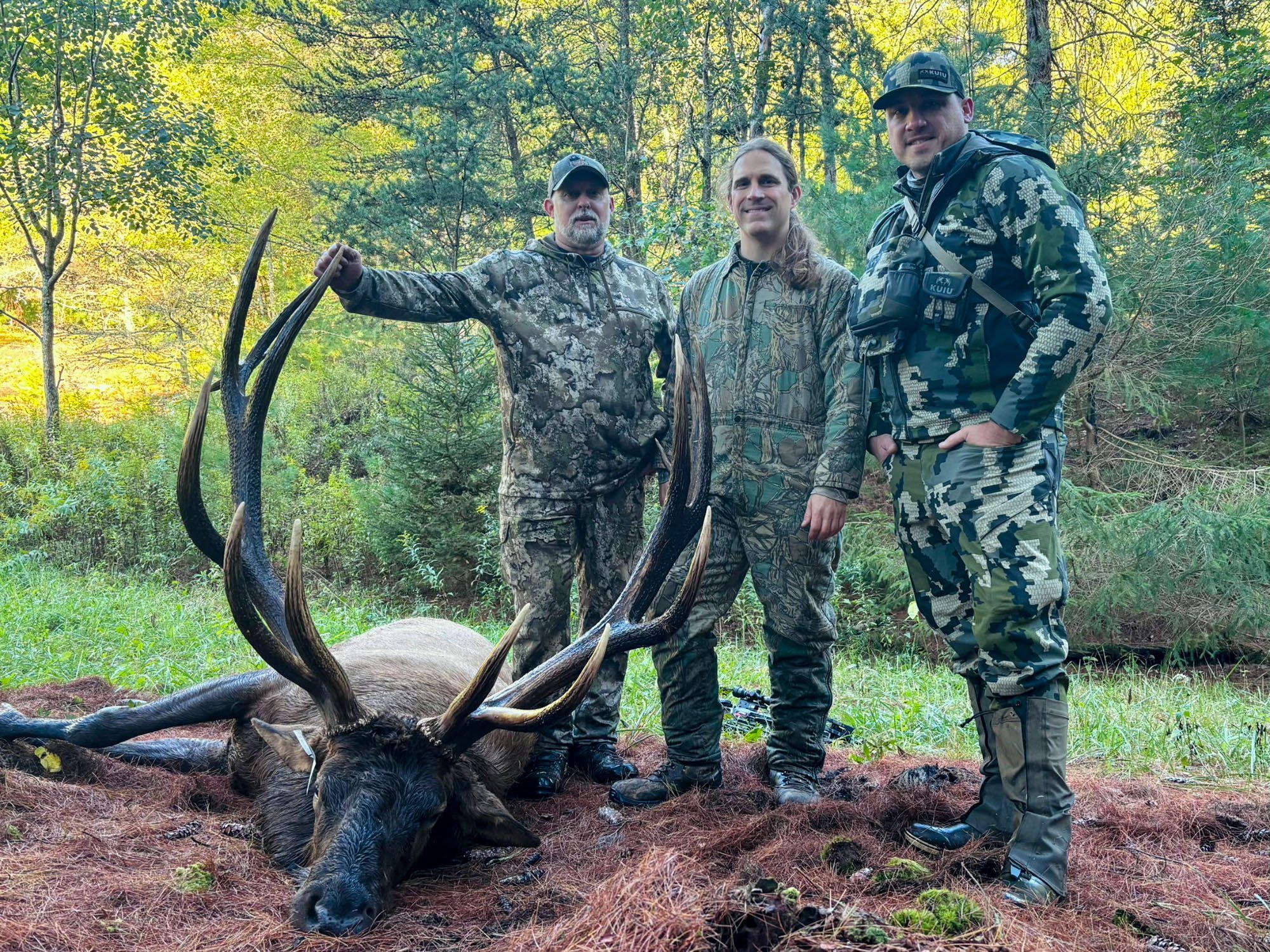 Three hunters in camo beside a big bull elk.