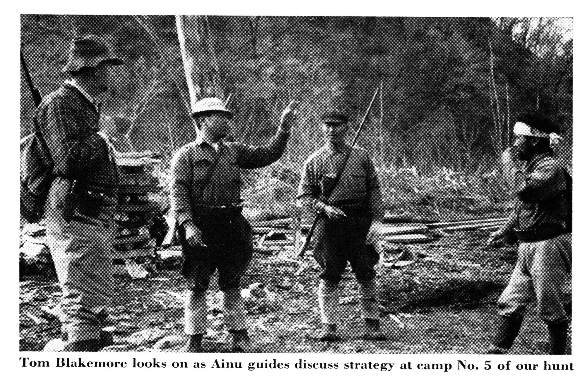 Four men in hunting clothes stand in a cleared forest camp; one gestures while the others listen, rifles slung over their shoulders.
