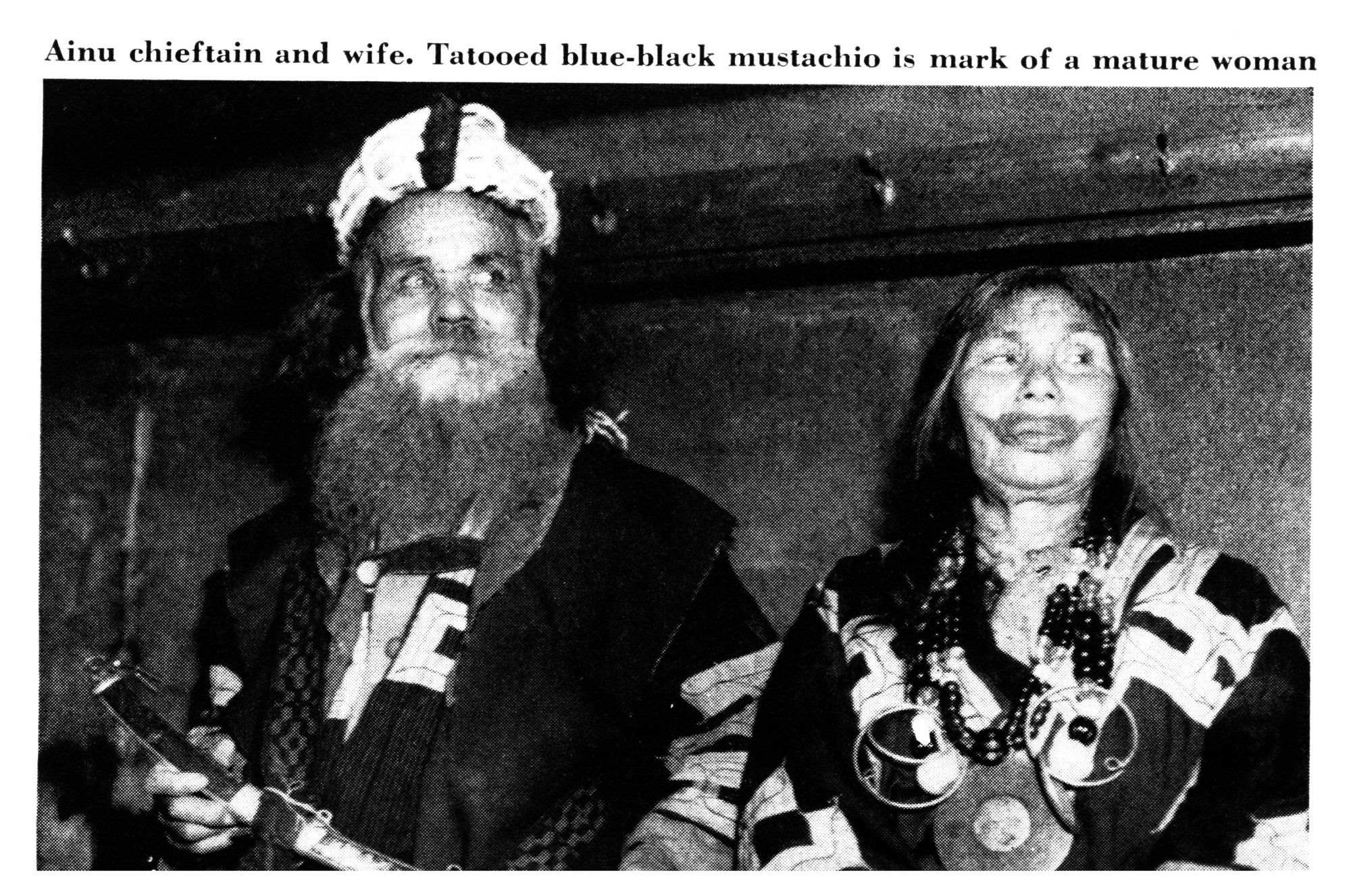 An Ainu chieftain with a long white beard stands beside his wife, who has a dark tattooed band around her mouth; both wear patterned robes and beaded necklaces.