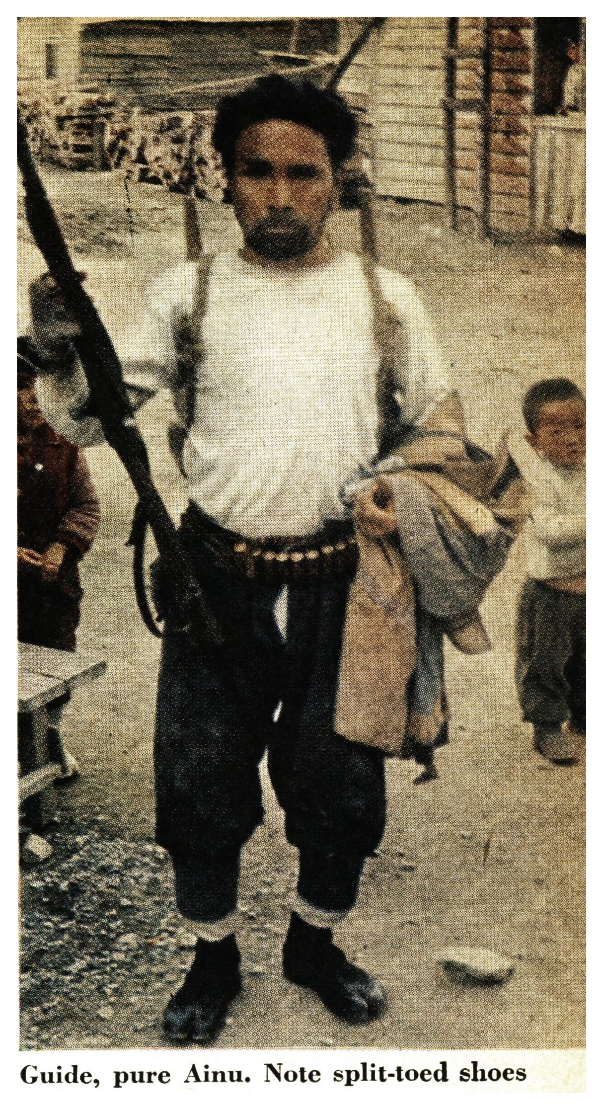 An Ainu hunting guide stands in a village street carrying a long-barreled shotgun over his shoulder, wearing split-toed shoes, with a child behind him.