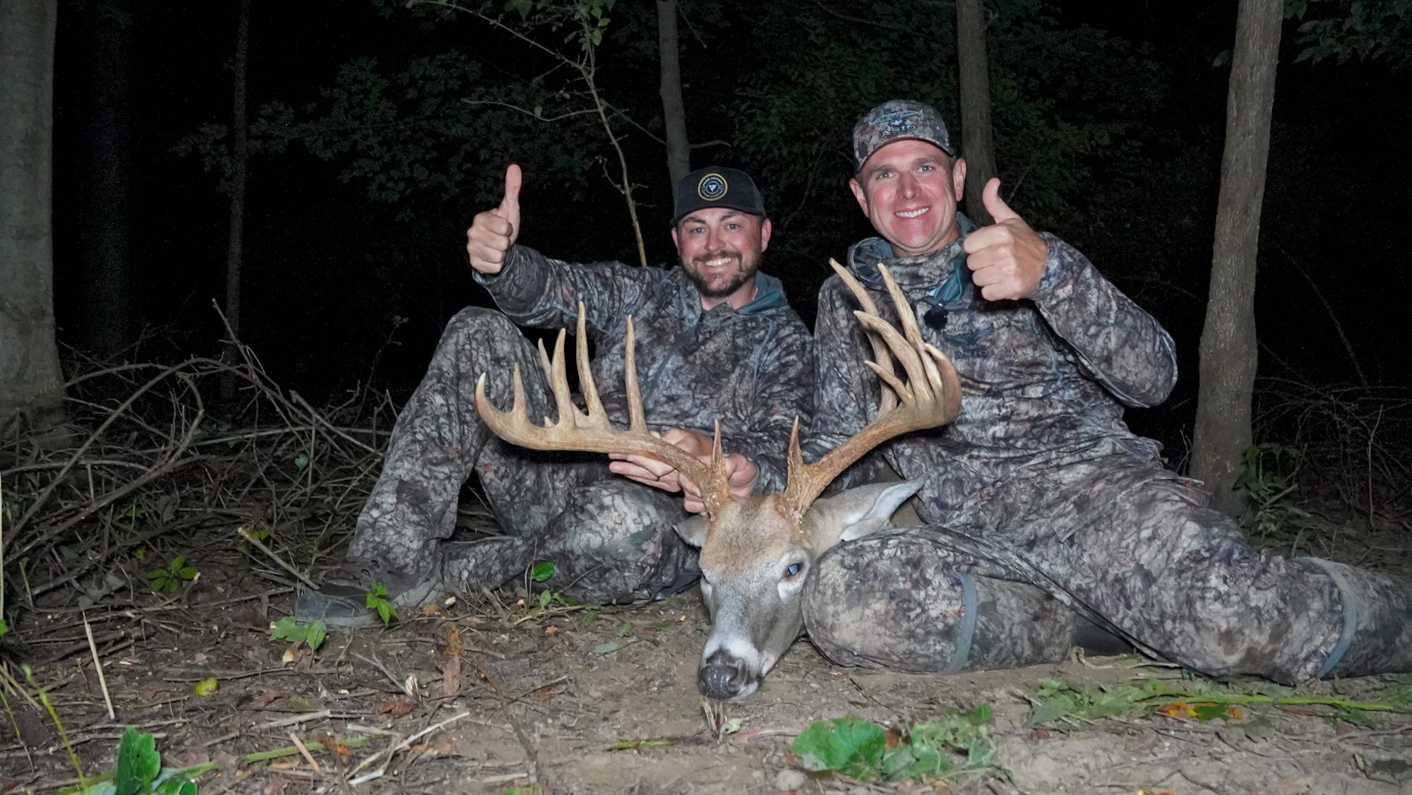 Two men in camo give thumbs up by a big buck.