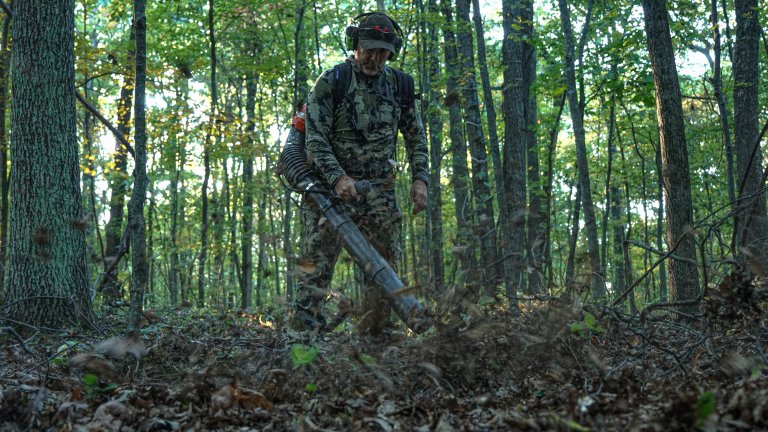A man in camo uses a backpack leaf blower in the woods
