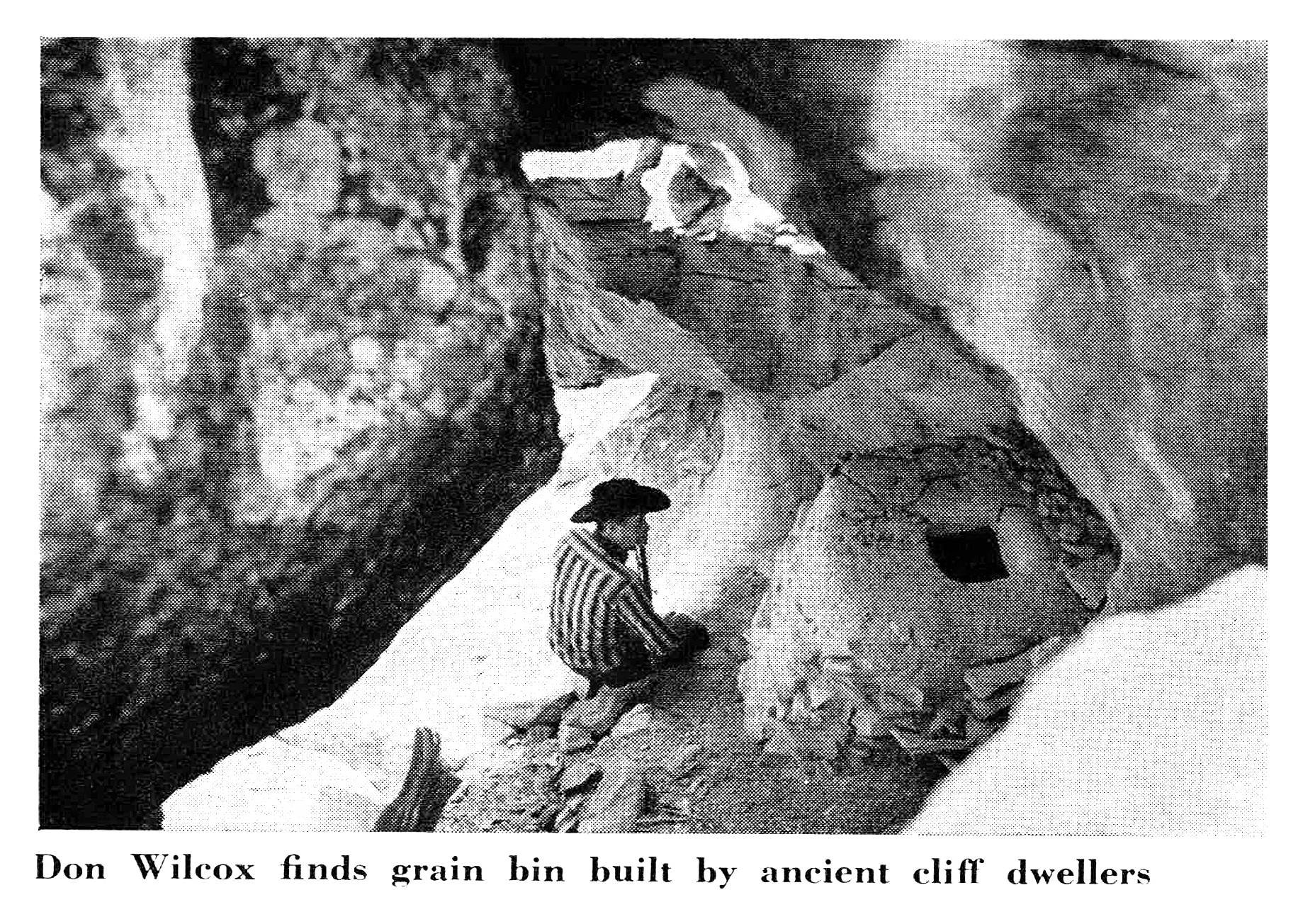 Black-and-white photo of a man in a cowboy hat crouching beside an ancient stone-and-mud granary built into a sandstone overhang.