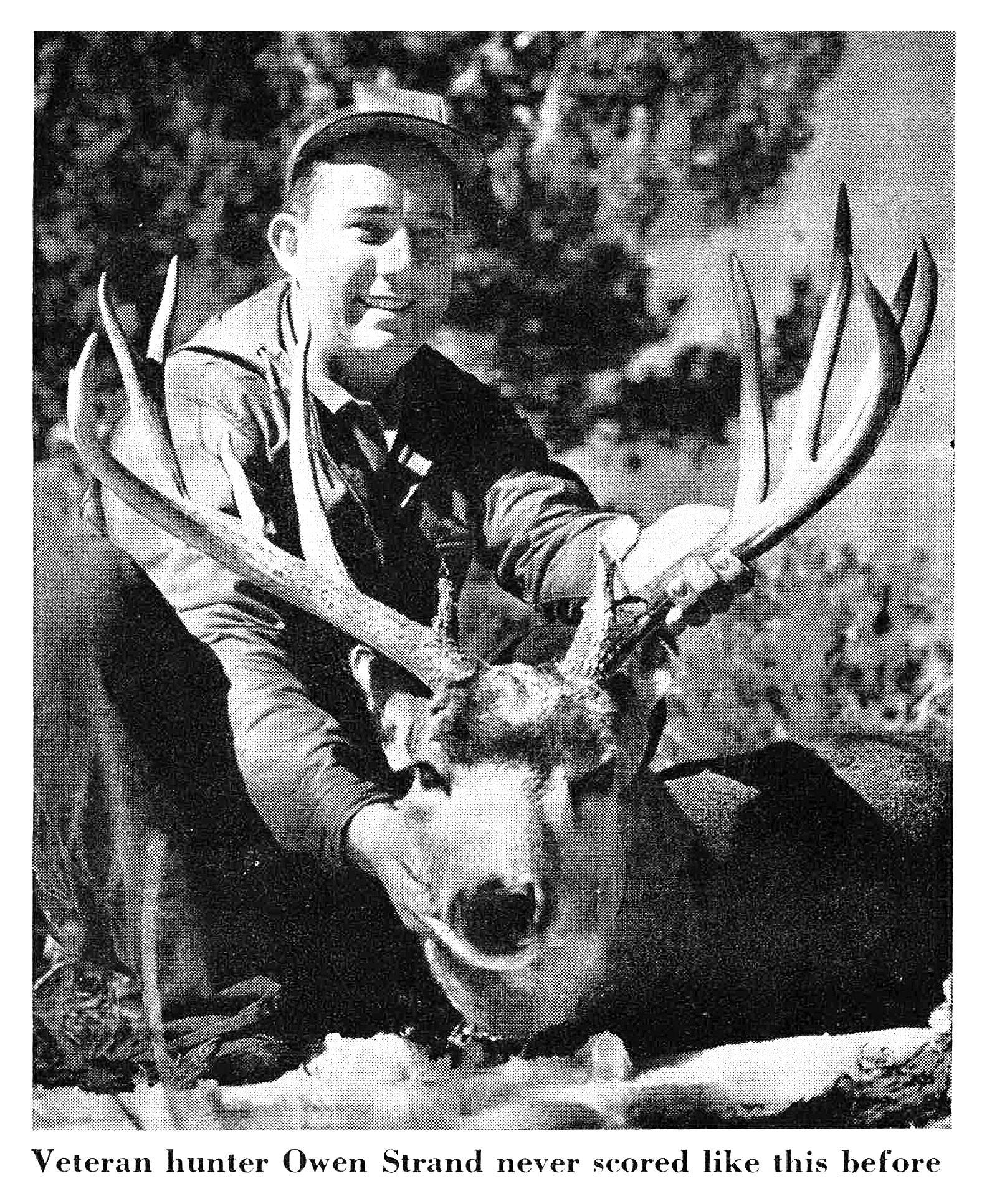 Black-and-white photo of a smiling hunter in a ball cap holding up the wide, tall antlers of a mule deer buck.
