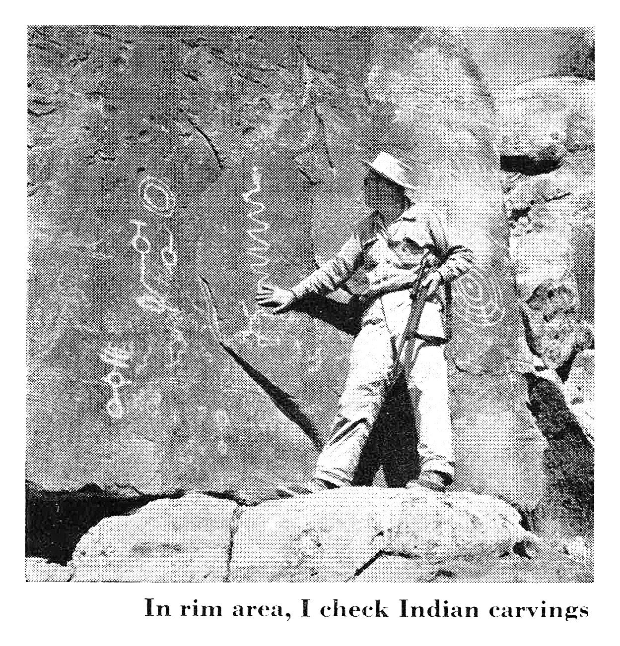 Black-and-white photo of a hunter with a rifle standing beside a rock wall covered in ancient petroglyphs.