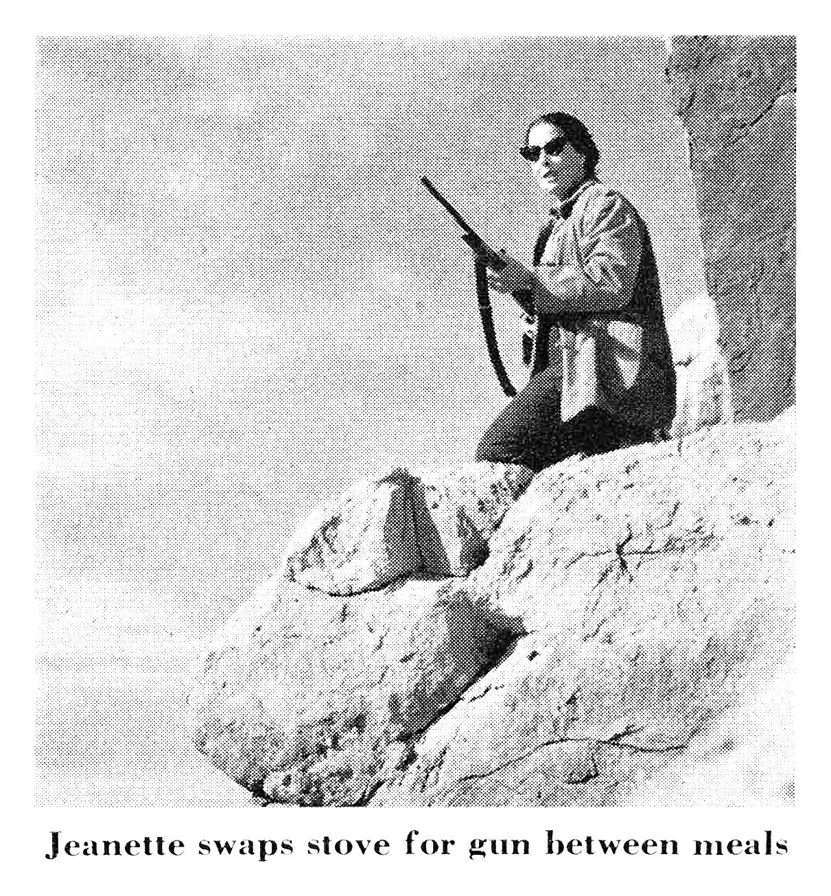 Black-and-white photo of a woman in sunglasses holding a rifle while standing atop a large boulder.