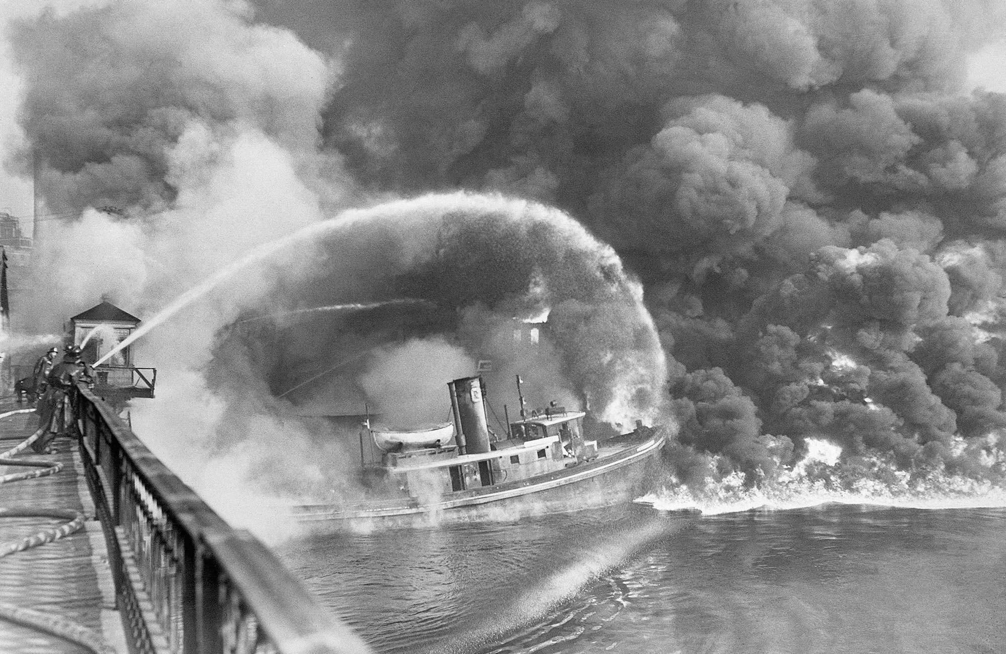 11/3/1952-Cleveland,OH- Firemen stand on a bridge over the Cuyahoga River to spray water on the tug Arizona, as a fire, started in an oil slick on the river, sweeps the docks at the Great Lakes Towing Company site in Cleveland Nov., 1st. The blaze destroyed three tugs, three buildings, and the ship repair yards.