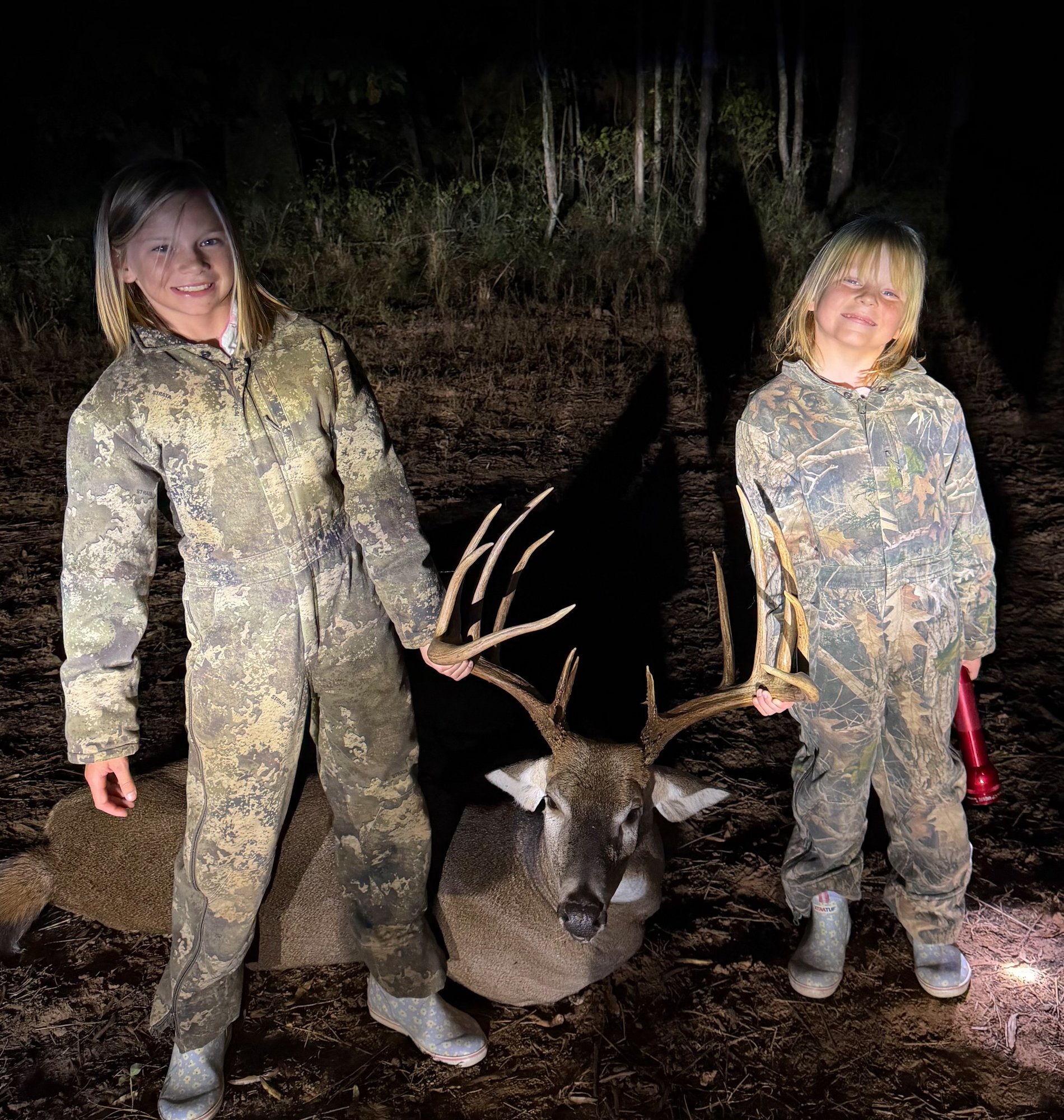 Two girls in camo suits hold the antlers of a big buck.
