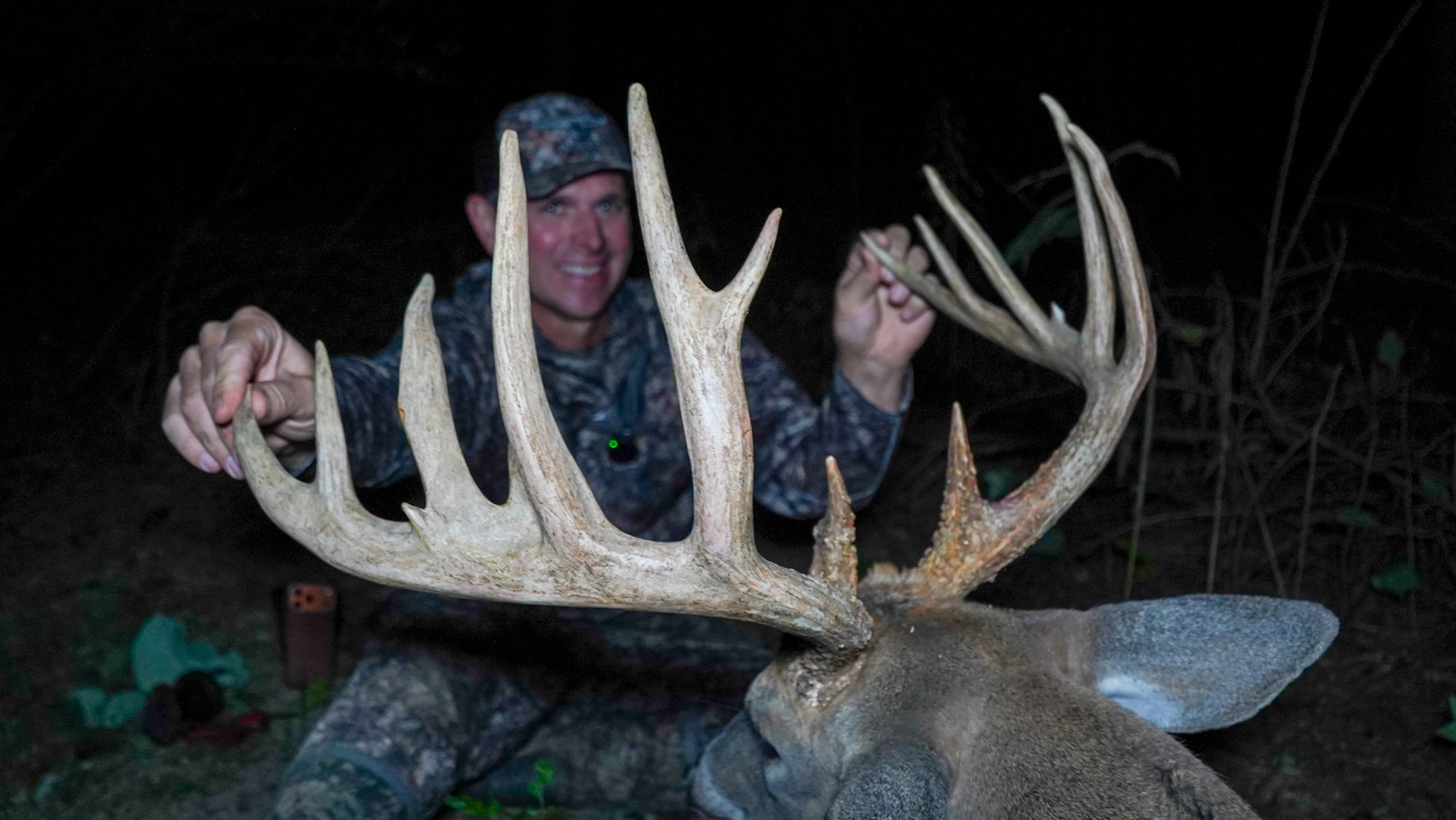 The antlers of a big buck fill the foreground of a nighttime pic while a hunter smiles in the background.