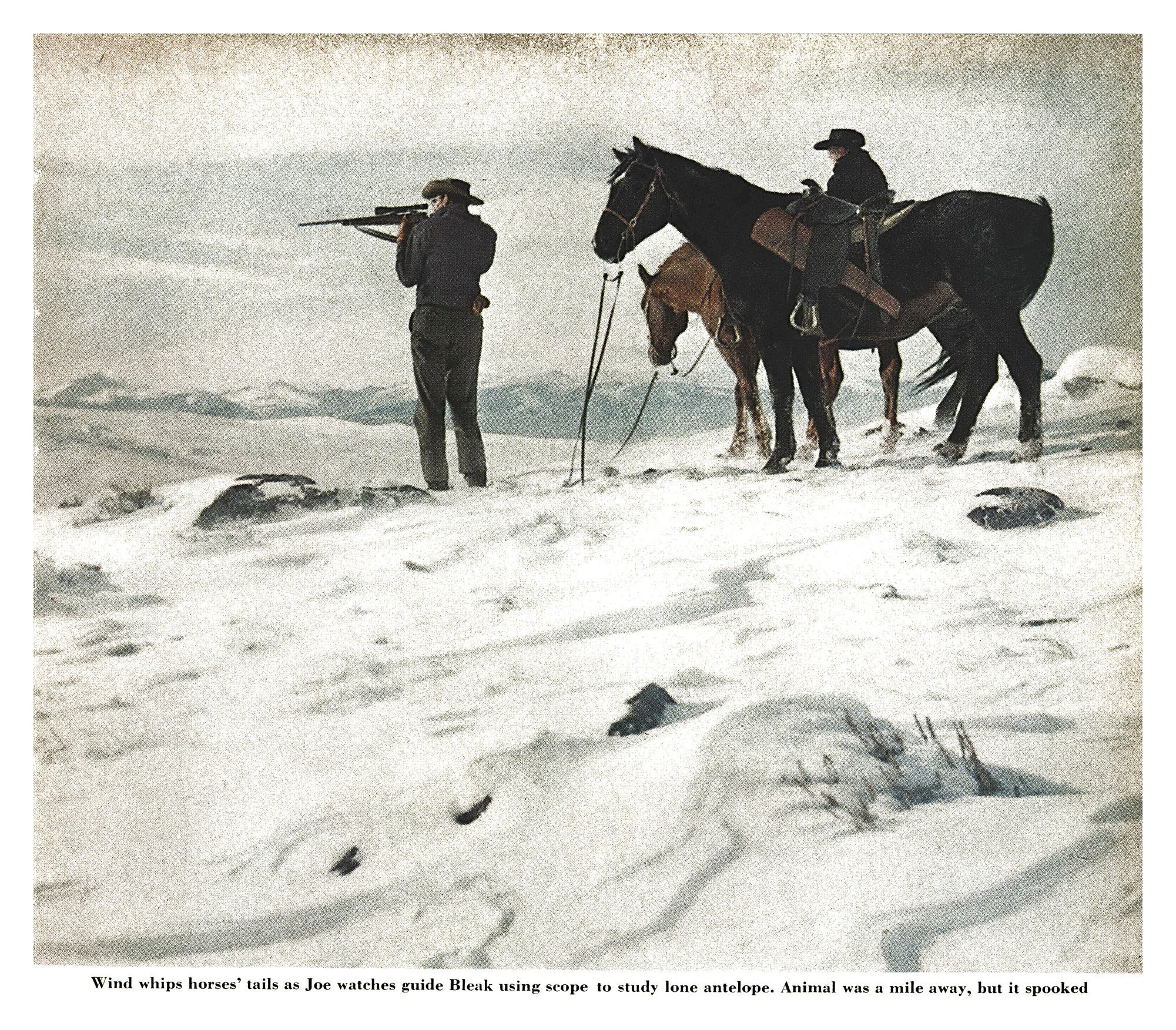A hunter aims a scoped rifle across a snowy, windblown ridge while a second rider sits on one of two horses standing beside him.