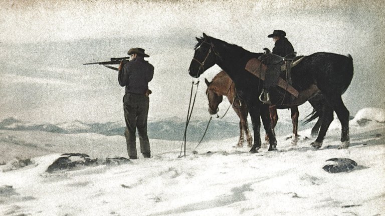 A hunter stands beside horses in the snow.