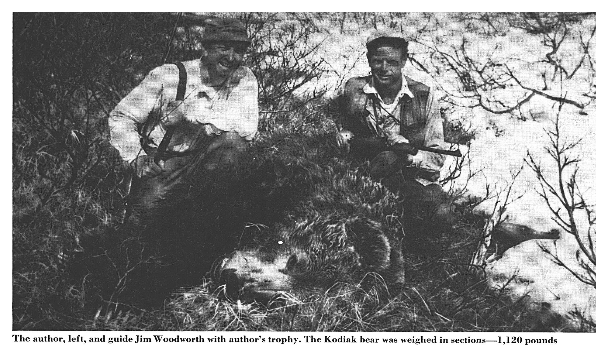 Black-and-white photo of two smiling hunters with rifles kneeling behind a huge Kodiak brown bear lying in grass and brush beside a snowbank.